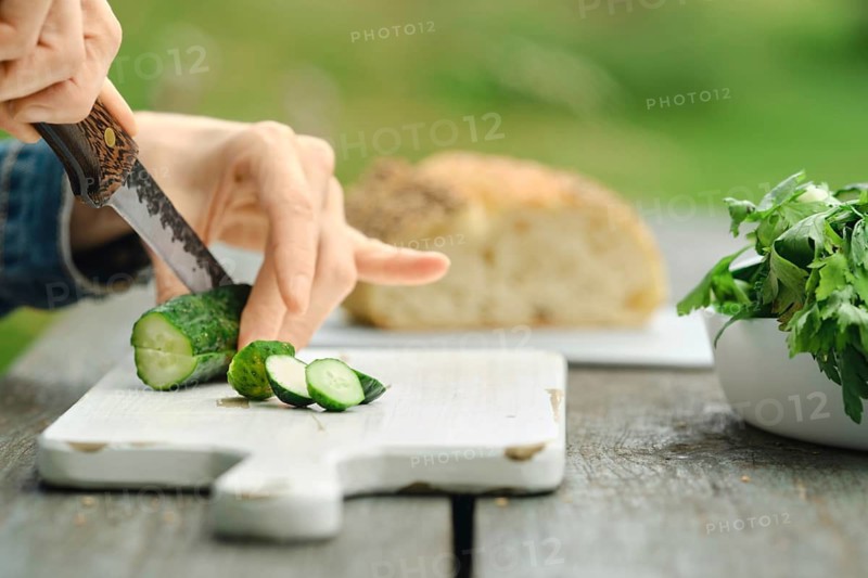 Hand holding knife cuts fresh cucumber on wooden board. Nearby, there is loaf of bread and bowl of herbs. This scene takes place in an outdoor setting during daylight