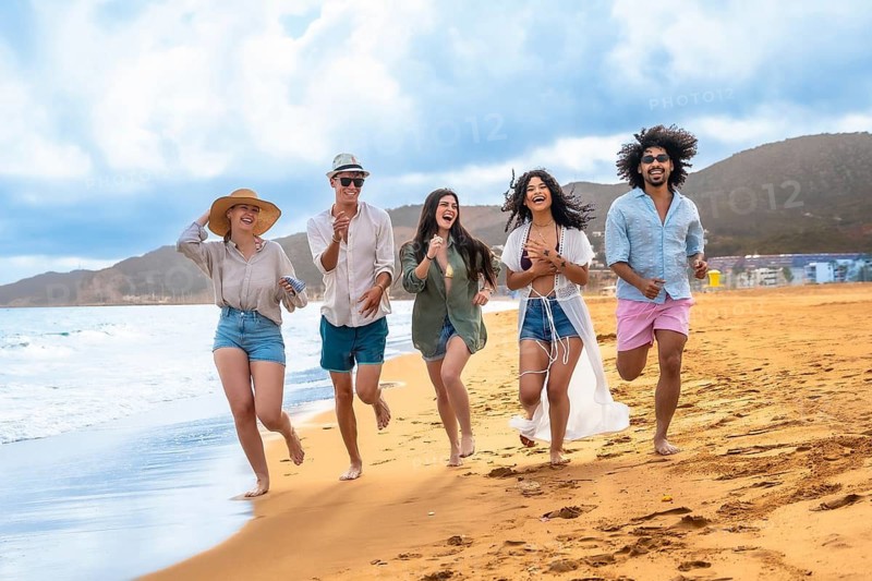 Diverse group of young adult friends running barefoot along the ocean shoreline on a sunny beach, celebrating summer vacation and enjoying collective happiness and freedom