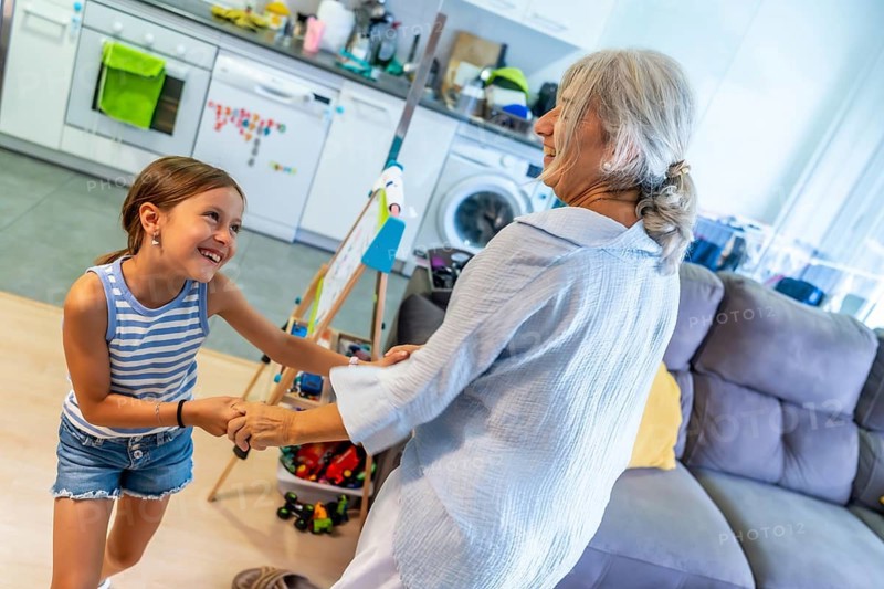 Happy grandmother and granddaughter holding hands and dancing together in a brightly lit home, laughing while enjoying generational family connection and playful activity