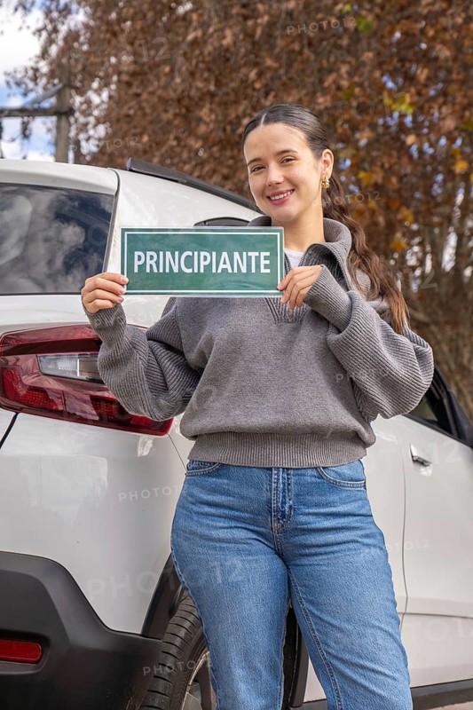 Smiling young woman holding green principiante beginner driver sign beside white SUV, outdoor autumn setting, new driver concept
