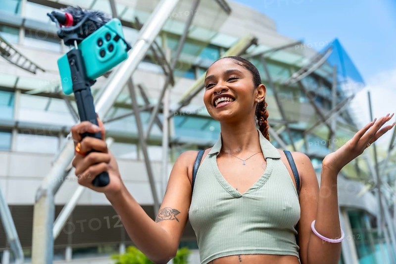 Young biracial woman actively recording content for her online followers, using a mobile phone with an attached microphone, smiling and gesturing while creating media in an urban setting