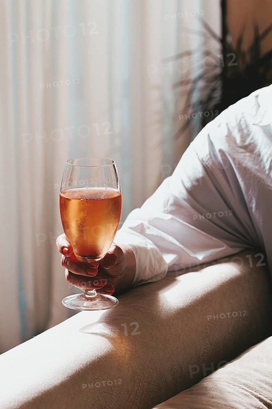 A person sits on a sofa holding a glass filled with a drink. Sunlight comes through the curtains, adding warmth to the scene. The setting looks inviting and cozy
