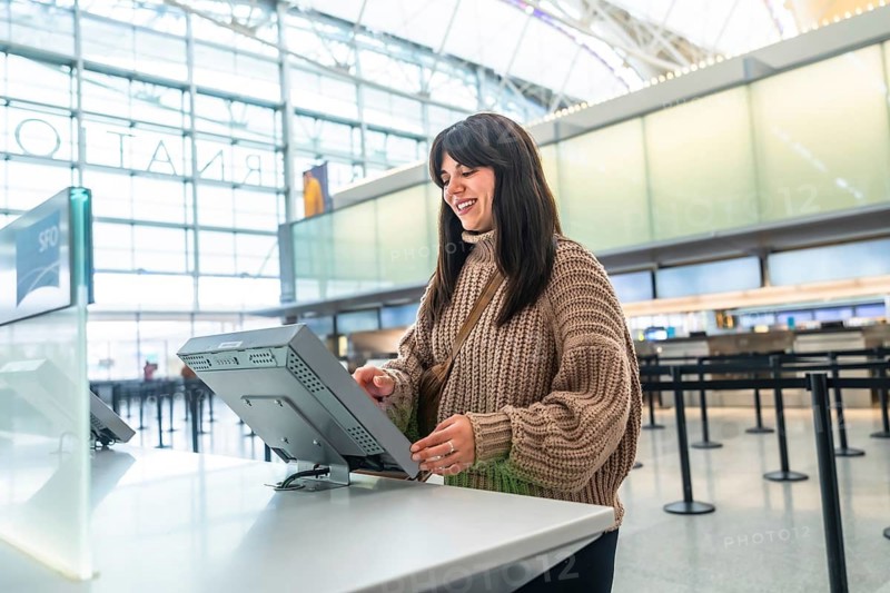 Young woman smiles while using a self service check in kiosk in a bright airport terminal, interacting with a touchscreen to manage her flight before departure
