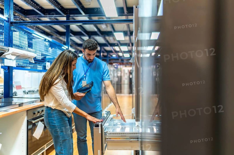 Sales assistant using a tablet to show product information to a female customer evaluating a refrigerator in a large contemporary home improvement store