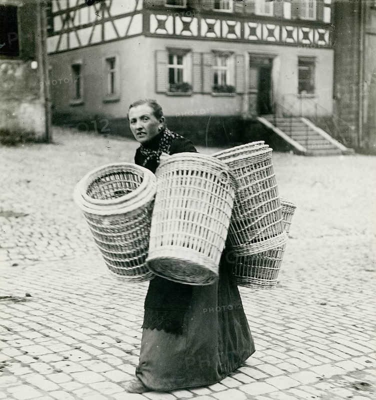 Bavaria, Franconia, woman's work, basket making, transport, poverty, home work, A woman carrying several large baskets across a paved street in front of a building, Philipp Kester, around 1906
