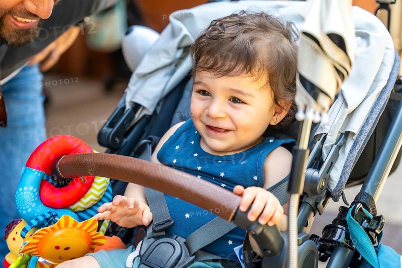 Happy baby girl in a star patterned top, safely strapped in a stroller with toys on the handle, smiling up at her father during a warm outdoor park stroll