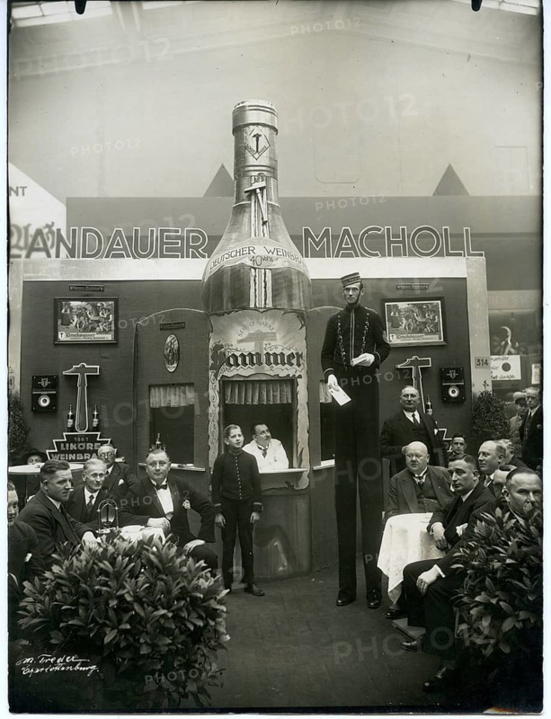 Trade exhibition, Berlin, Landauer Macholl stand from Heilbronn, brandy hammer, advertisement, Jewish, 1938 Aryanized, people gather around a large bottle advertisement at a trade fair with decorative backdrop, 1932