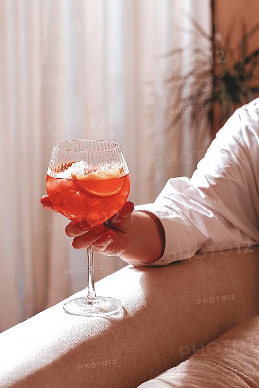 A person is sitting on a couch in a bright room. They hold a glass filled with a red drink and ice. Sunlight comes in through a nearby window