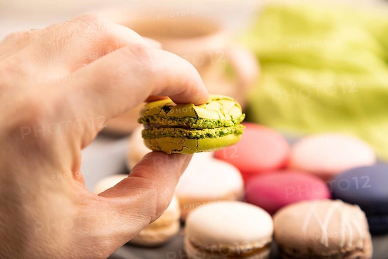 Macarons colourful cookies dessert with hand on brown concrete background with green linen textile, close up, side view, selective focus