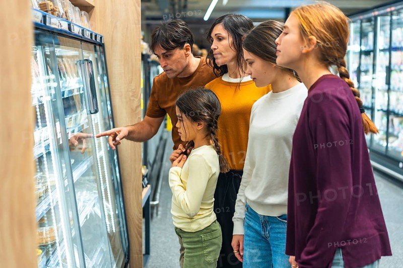 Family group of mother, father, and three daughters standing next to a refrigerated display in a grocery store, with the father pointing at items inside while looking for products