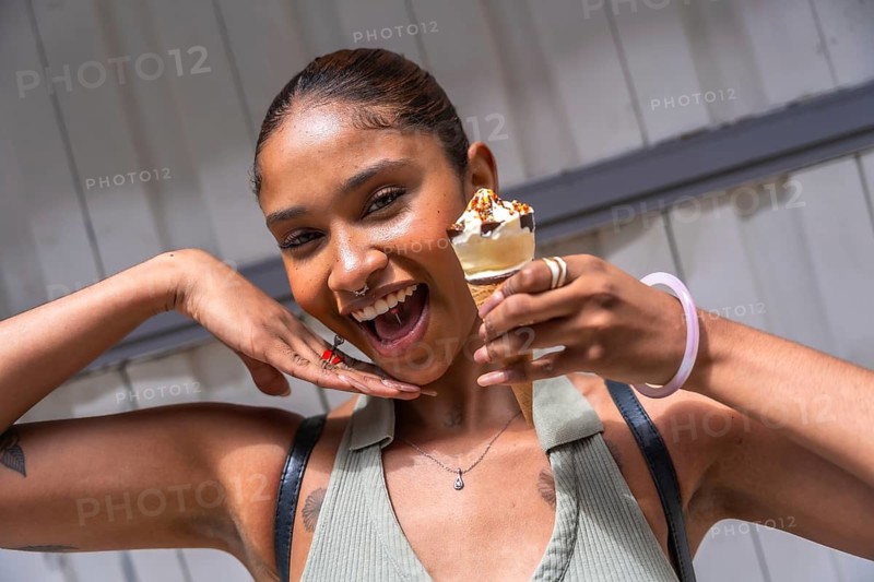 Young woman with radiant skin smiling brightly, holding a delicious ice cream cone with chocolate syrup and nuts, expressing sheer joy and embracing summer vibes