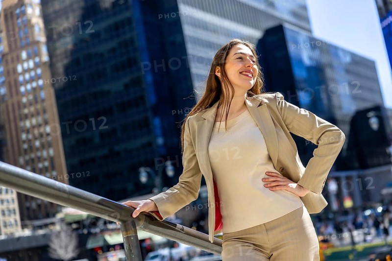 Professional businesswoman smiling confidently, leaning on a railing in manhattan's financial district, looking away toward future possibilities amid skyscrapers and sunny streets