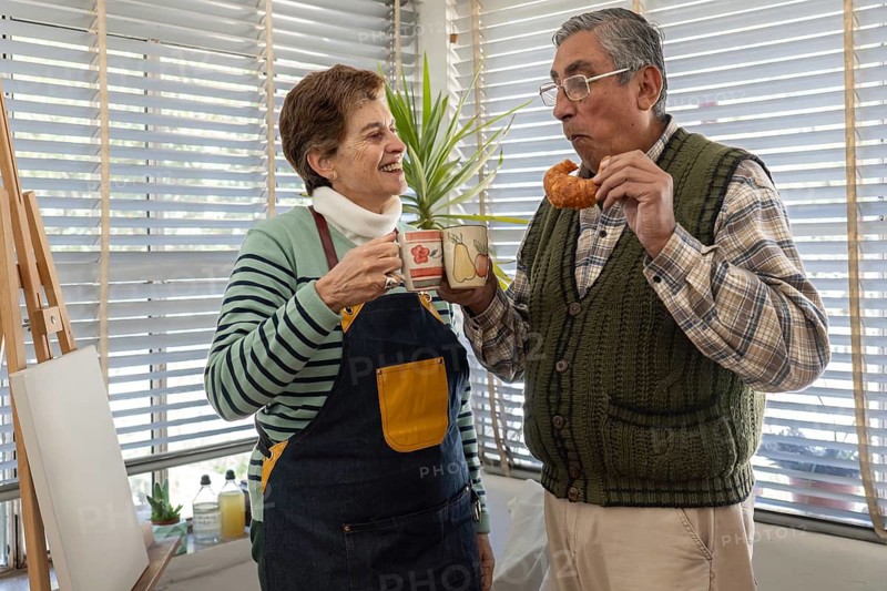 A senior woman in a denim apron passes a coffee mug to her husband who is eating a croissant, both standing together and smiling in a light-filled home space near an easel and indoor plants