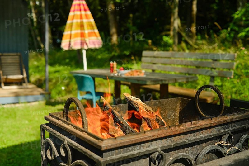 Fire pit burns wood in backyard setting. Green grass surrounds fire pit. Nearby table with drinks. Umbrella provides shade