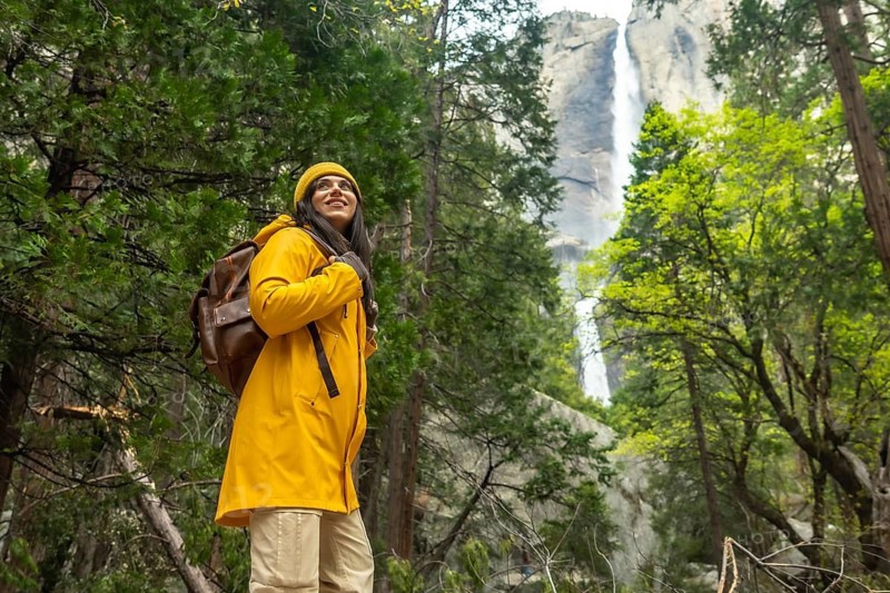 Woman in a yellow raincoat and backpack hiking through the forest, looking up with a happy expression, enjoying the view of lower yosemite falls and the surrounding nature