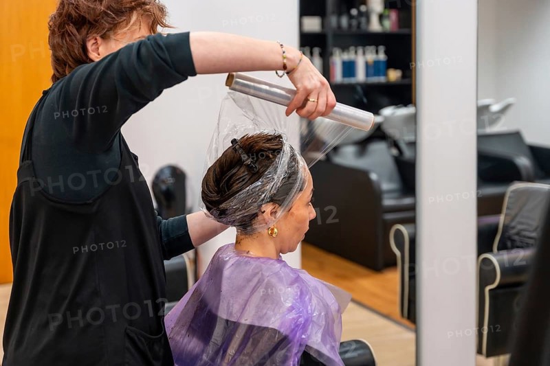 Hairdresser applying plastic wrap over a client's hair after a coloring treatment, working inside a modern beauty salon focusing on hair care and professional services