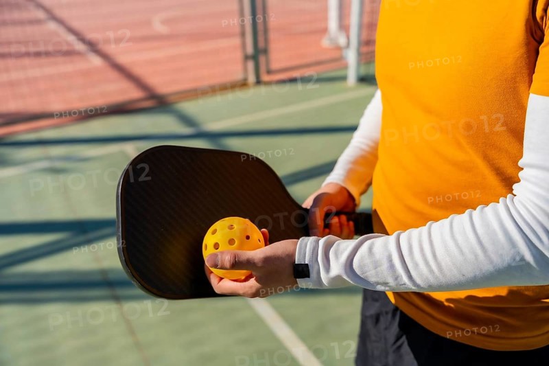 Player prepares to serve during a pickleball game, holding a black carbon paddle and bright yellow perforated ball, standing on an outdoor sports court