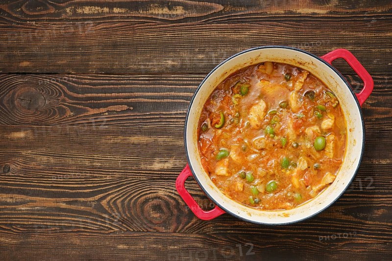 Pot filled with stew sits on wooden table. Stew has meat and green vegetables mixed together. Food is warm and ready to eat