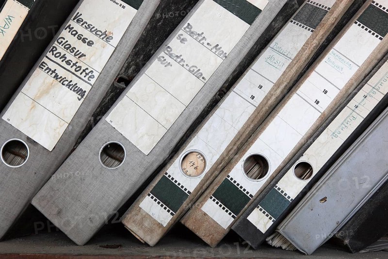 Office scene around 1982, lost place, abandoned location, classic file folders on a wooden shelf, some of the folders are labelled in detail by hand and provide information about their contents, mixed up, dirty