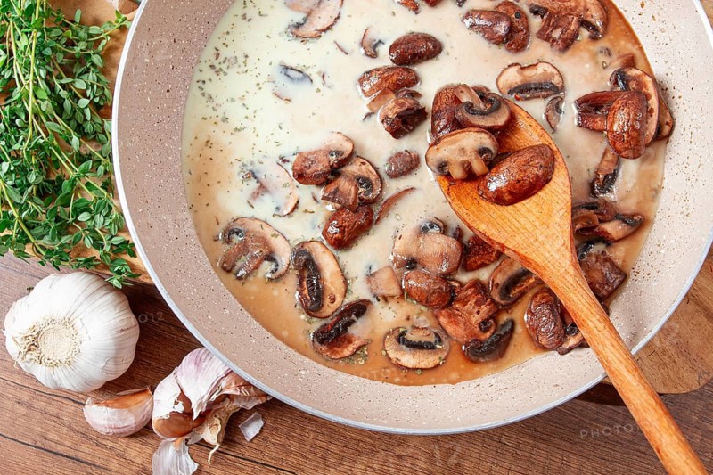Mushrooms are cooking in a creamy sauce with garlic and herbs in a pan. Fresh thyme and garlic are on the side. The setting is a wooden table with natural colors