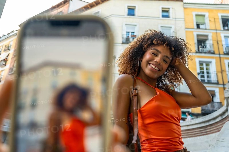 Young african american woman with curly hair smiling directly at the camera, posing for a selfie using a smartphone on a city street during summer travels