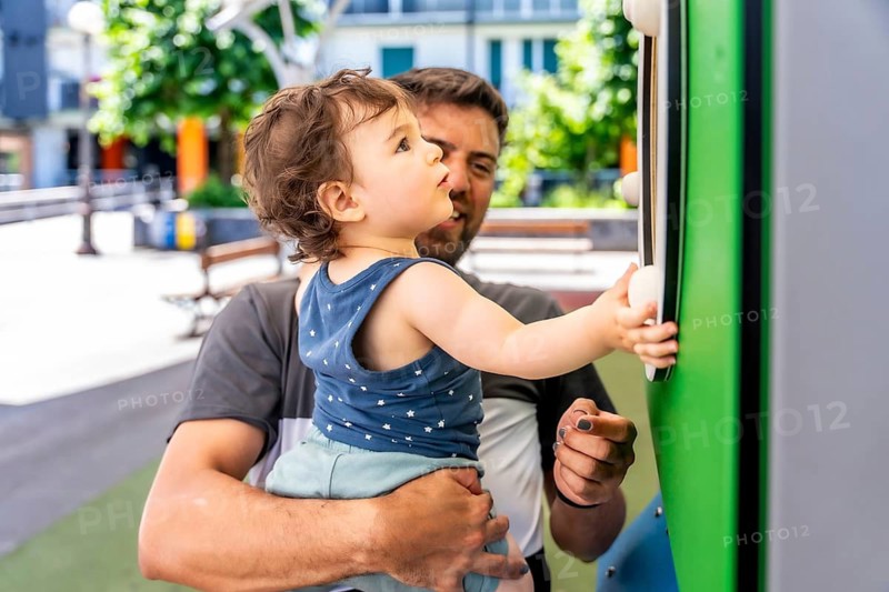 Caring father holding a young child, who is exploring and interacting with a colorful sensory play panel at a city park playground on a beautiful sunny day