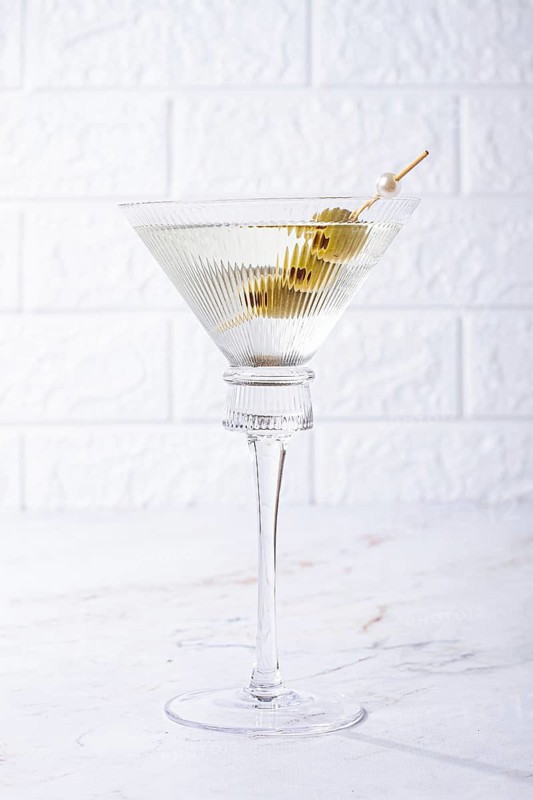A clear glass cocktail sits on a white marble surface. Three green olives are placed on a skewer resting inside the glass. The light shines on the glass, highlighting its shape