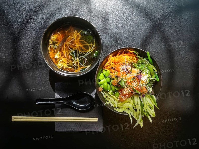 Lunch in Asian style. Shiro miso soup and poke bowl with salmon, cucumbers, green beans, carrots and chukka salad. A neatly arranged meal with two bowls placed side by side on a dark surface. One bowl contains clear noodle soup with vegetables, while the other features a colorful poke bowl with salmon, edamame, carrots, cucumber, seaweed and sesame seeds. A black spoon on a napkin and a pair of chopsticks complete the modern healthy food presentation