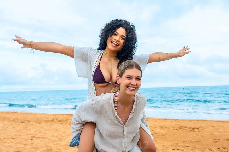 Two cheerful young women celebrating friendship and fun with a piggyback ride on a sandy beach, embracing joy and freedom during their warm weather vacation