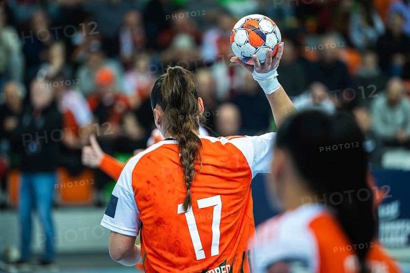 Female handball player wearing an orange jersey with number 17 holding the ball, ready to throw during a professional match in a packed arena in Lubin, Poland