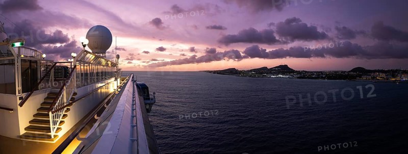 Panoramic view from a Caribbean cruise ship deck at sunset showing a purple sky over the island coastline of Willemstad Curacao with the ocean at dusk