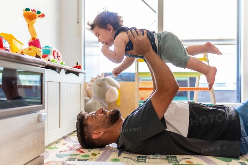 Father lying on a colorful playmat, joyfully lifting and playing with his smiling baby son in an airplane game, fostering a strong bond and creating happy family moments