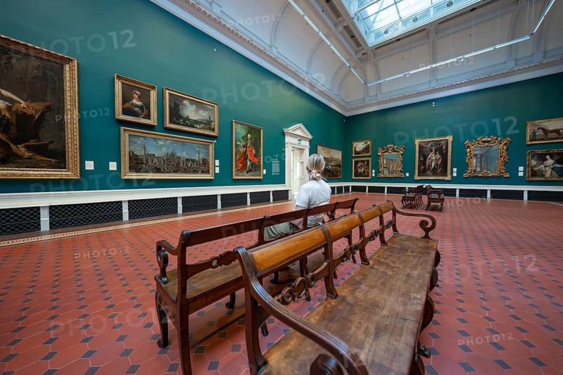 A visitor looks at paintings in an ornate gallery with green walls and wooden benches, National Gallery of Ireland, Dublin, Ireland