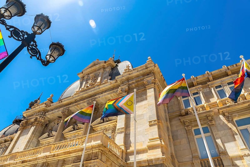 Cartagena city hall building, known as palacio consistorial, displaying rainbow and progress pride flags, symbolizing lgbt rights and inclusion, under a bright blue sky in murcia, spain