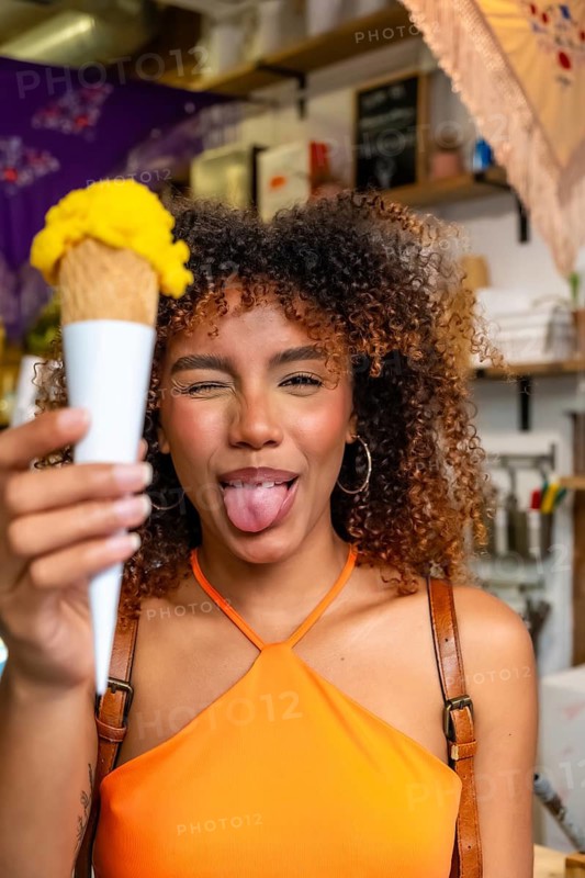 Young curly haired woman holding a vibrant yellow ice cream cone, winking and sticking out her tongue, showing a cheerful and playful mood in a sweet shop