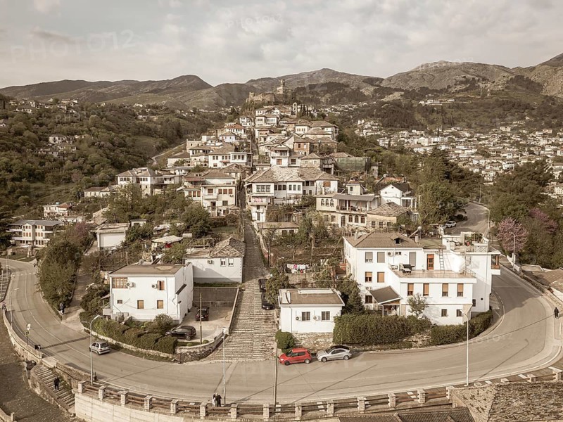 Aerial panoramic view massive stone Gjirokaster Castle on hilltop, surrounded by historic Stone City buildings - Unesco world heritage site. Rugged peaks of Gjere Mountains in Albania. Sepia filter