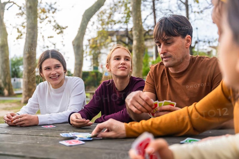 Father and two daughters play cards at a park picnic table, enjoying a relaxed, sunny day of bonding, laughter and casual family fun in nature