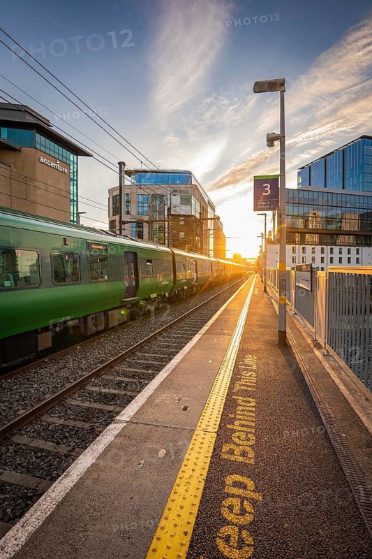 A green train stops at a platform as the sun sets between modern buildings, Dockland, Dublin, Ireland