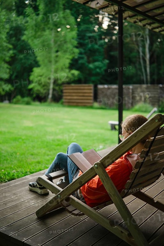 Woman sits on lounge chair in backyard surrounded by grass and trees reading book in afternoon. Calm and quiet atmosphere enhances the enjoyment