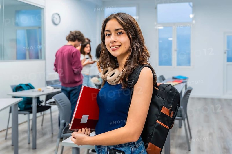 Young woman student smiling at camera, carrying a backpack and a notebook, standing in a brightly lit modern classroom with other students in the background