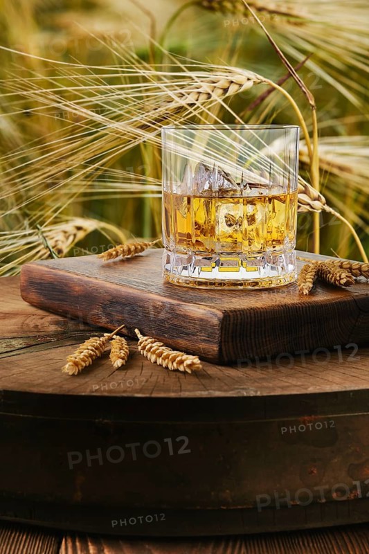 Whiskey glass on wooden surface beside wheat stalks. The background shows wheat field under bright sunlight