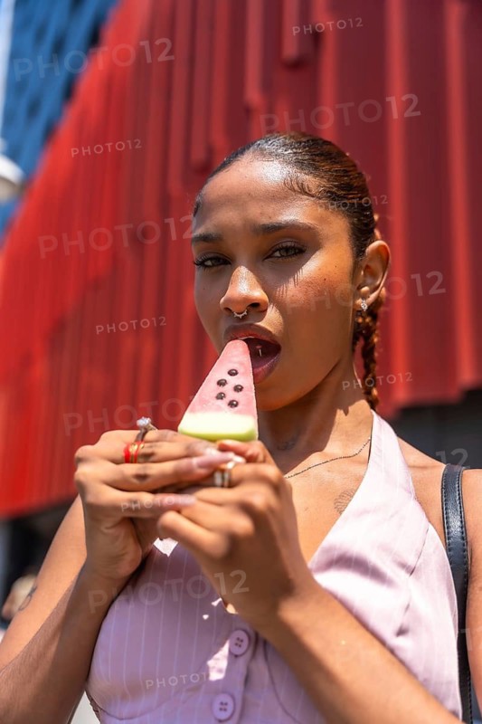 Young woman refreshing with a watermelon shaped popsicle on a hot summer day, holding the frozen treat near her mouth in an urban setting