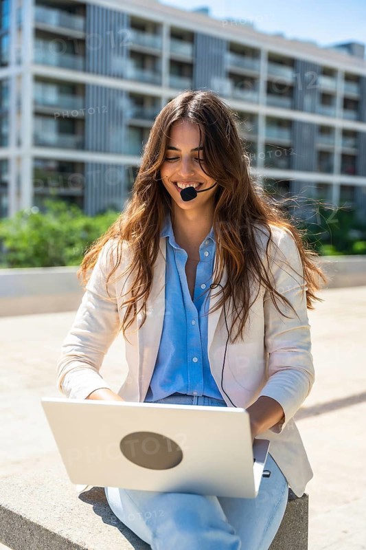 Smiling businesswoman wearing a headset, working remotely with her laptop in an urban outdoor setting, embodying modern communication, connectivity, and the freedom of flexible work