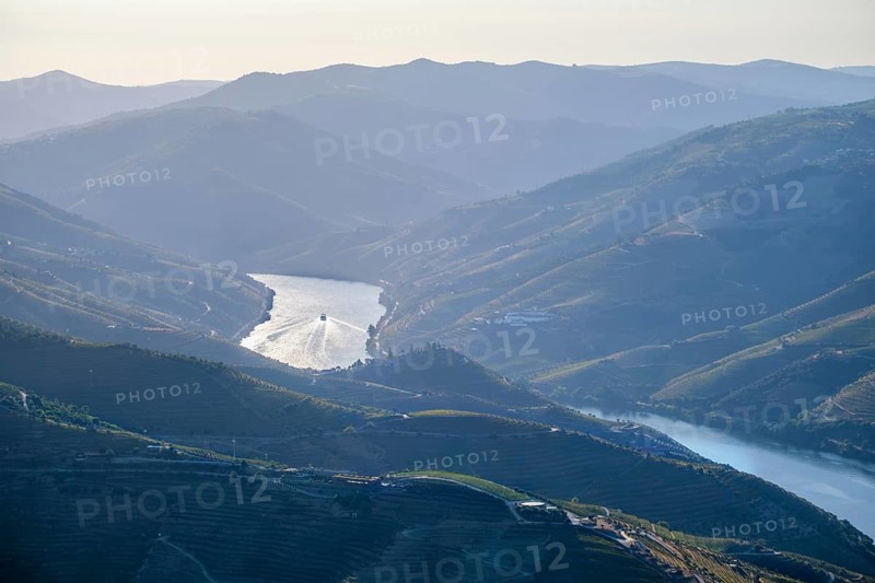 The drone view captures sunrise over the Douro Valley river corridor in Portugal. Vineyard hills surround the area, with a tourist boat moving along the water