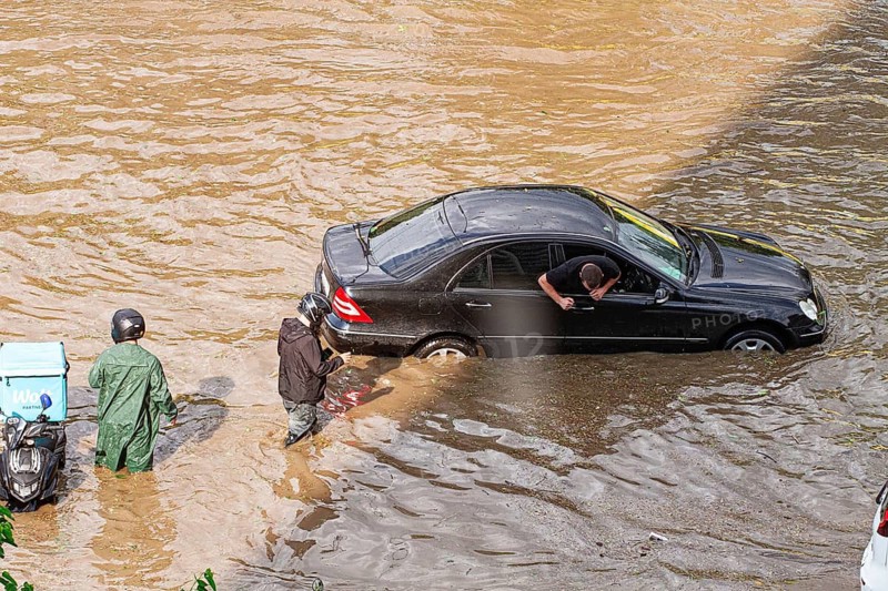 In a city affected by heavy rainfall, cars are submerged in water, and people are seen wading through the flooded streets. The scene shows the impact of severe weather conditions