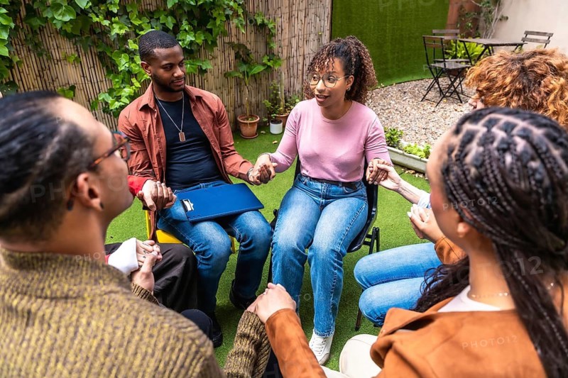 Diverse people participating in an outdoor group therapy session, sitting in a circle, holding hands, and sharing experiences, symbolizing support, connection, and mental health awareness
