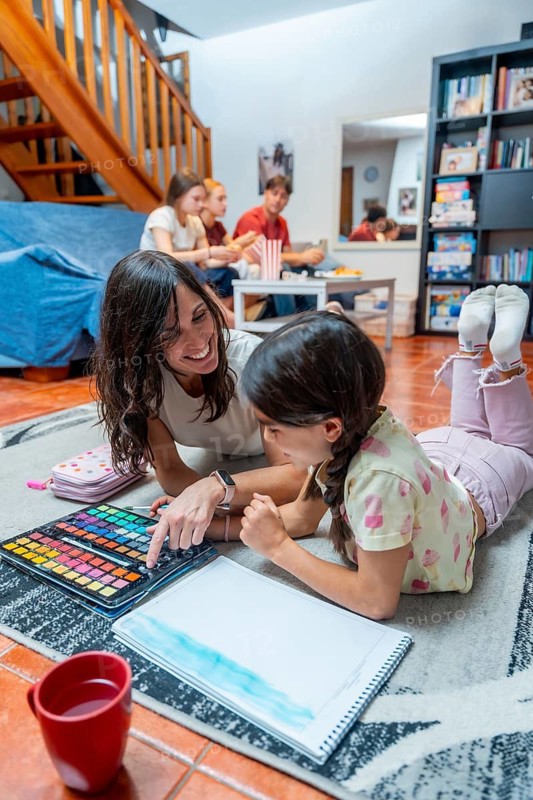 Mother and daughter paint with watercolors on the living room floor, smiling and bonding over creative play while family relaxes nearby in a cozy home setting