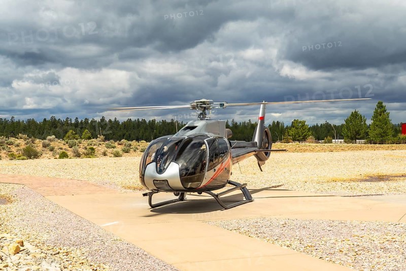 Helicopter parked on an asphalt helipad, preparing for scenic flying tours, with a dark cloudy sky overhead and desert scrub and pine trees in the background