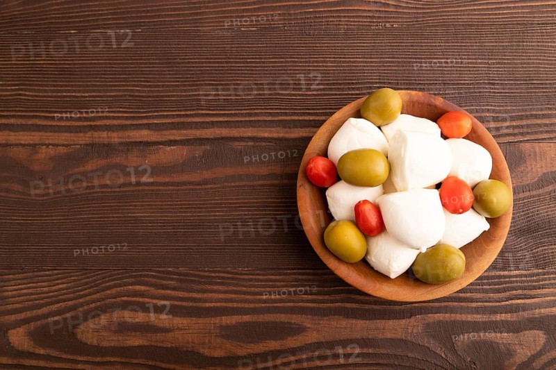 White Mozzarella cheese, with tomatoes and olives in wooden bowl on brown wooden background, top view, flat lay, copy space