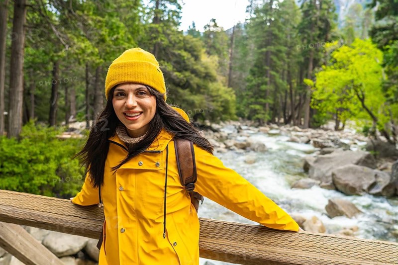 Woman standing on a wooden bridge, smiling confidently while enjoying the fresh air and natural beauty of yosemite national park, with a flowing river and lush forest in the background
